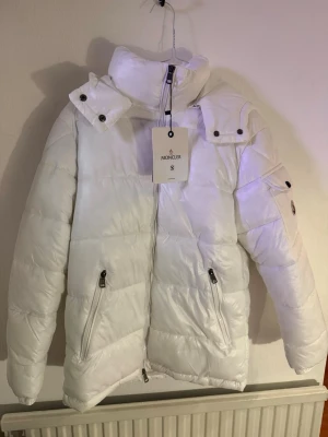 Vit dunjacka från Moncler med huva - Säljer en snygg vit dunjacka från Moncler i storlek S. Jackan har avtagbar huva, dragkedja framtill, två fickor med dragkedja och en ficka med knapp på, scannar man qr koden kommer man in på hemsidan samt att loggan på armen tar sig också in på hemsidan, kan dock inte svara på om den är äkta