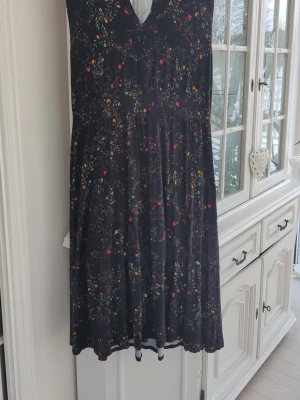 Svart blommig klänning - Svart klänning med  blommönster i rött, blått, orange och grönt. Klänningen har v-ringning och är ärmlös, med en lätt utställd kjol. Öppen detalj i ryggen. 