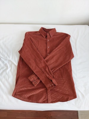 Orange / Rostbrun manchesterskjorta - Säljer en snygg rostbrun skjorta i manchester med klassisk krage och knappar framtill. Skjortan är långärmad och har en avslappnad passform som passar perfekt till jeans eller chinos. Materialet är mjukt och ger en retrovibe.