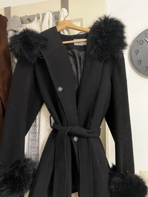 Hollies kappa med fuskpäls  - Stilren svart kappa med luva och fuskpäls på huva och ärmslut. Knytskärp i midjan ger en snygg siluett. Kappan är fodrad och har knappar framtill. Perfekt för dig som vill ha en elegant och trendig look under kalla dagar. Köpt för 1800kr OBS liten i storlek