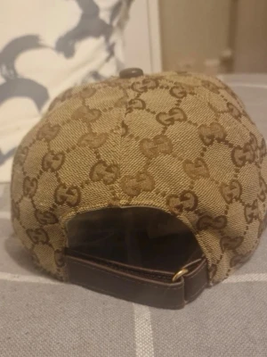 Beige keps med GG-mönster  - Säljer en beige keps från Gucci med det klassiska GG-monogrammet i brunt över hela kepsen. Justerbar rem i brunt läder baktill och detaljer i canvas. Perfekt för dig som vill ha en lyxig och tidlös accessoar till din outfit.