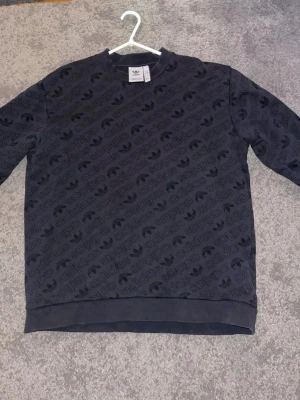 Svart Adidas sweatshirt med logga - Svart sweatshirt från Adidas med all-over tryck av Adidas-loggan och treklöver. Tröjan har rund hals, ribbade muddar och är gjord i mjuk bomullsblandning. Perfekt för dig som gillar streetwear och vill ha en skön och stilren look.