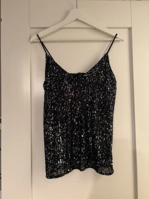 Svart paljettlinnetopp - Glittrig svart linne med tunna axelband och täckt av paljetter. Perfekt för dig som vill sticka ut och glänsa. Snyggt att styla med jeans eller kjol för en trendig look.