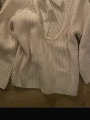 Vit ribbstickad tröja med rosett - Säljer en vit ribbstickad tröja med lång ärm och rund halsringning. Tröjan har en snygg rosettdetalj i nacken och är gjord i ett mjukt, stretchigt material. Perfekt för dig som gillar stilrena och enkla plagg med en twist.