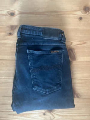 Mörkblå jeans från Nudie Jeans - Säljer ett par mörkblå jeans från Nudie Jeans med klassisk femficksdesign och snygga detaljer på bakfickan. Jeansen har en rak passform och är tillverkade i mjukt denimtyg. Perfekta för dig som gillar stilrena och tidlösa jeans.