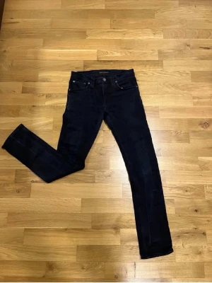 Svarta slim fit jeans från Nudie - Säljer ett par svarta slim fit jeans från Nudie Jeans med klassisk femficksmodell och raka ben. Jeansen har diskreta sömmar och är tillverkade i stretchigt denimtyg för extra komfort. Perfekta för en clean och stilren look.