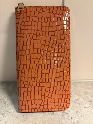 Orange plånbok med krokodilstruktur - Snygg orange plånbok i konstläder med glansig krokodilpräglad yta. Plånboken har guldfärgad dragkedja och matchande handledsrem. Perfekt för dig som vill ha en trendig accessoar med plats för kort och kontanter.