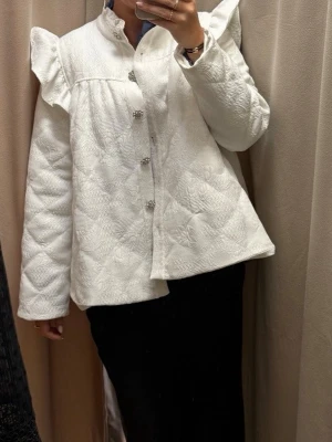 custommade jacka - Stilren vit quiltad jacka med volangdetaljer på axlarna och dekorativa knappar framtill. Från custommade. Köpt för 3500kr. Obs: vet ej storlek men passar S-L. 