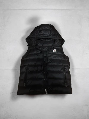 Svart dunväst från Moncler med luva - Snygg svart dunväst från Moncler luva och dragkedja framtill. Västen har två praktiska fickor med dragkedja och den klassiska Moncler-loggan på bröstet. Perfekt för lager på lager och streetstyle.