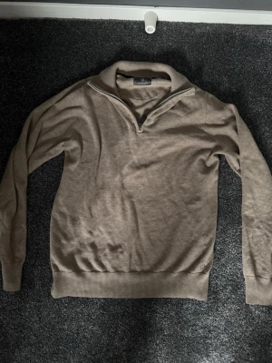 Beige halvzip tröja från Oscar Jacobson - Säljer en stilren beige tröja från Oscar Jacobson med halvzip och hög krage. Tröjan har långa ärmar, ribbade muddar och nederkant. Perfekt för dig som gillar en clean och klassisk look.