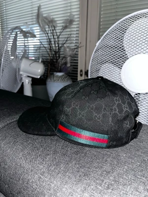 Svart Gucci keps med GG-mönster - Säljer en svart keps från Gucci med det klassiska GG-monogrammet över hela kepsen. På sidan finns en grön och röd rand som ger en lyxig touch. Justerbar rem bak för perfekt passform. Materialet känns slitstarkt och exklusivt.