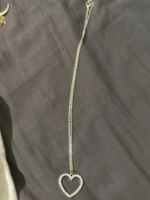Silverfärgat halsband med hjärthänge - Snyggt halsband i silverfärg med ett stort hjärthänge dekorerat med små gnistrande stenar. Kedjan är tunn och elegant, perfekt för att ge en romantisk touch till din outfit. Hjärtat har en öppen design och ger ett stilrent intryck.