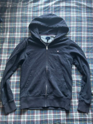 Mörkblå zip hoodie från GANT - Säljer en mörkblå hoodie från GANT i storlek S. Hoodien har dragkedja framtill, två fickor och en broderad GANT-logga på bröstet. Insidan av huvan är grå och har en snygg randig detalj. Perfekt för en chill och stilren look.