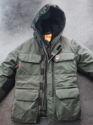 Fjällräven Hydratic vinterjacka - Säljer en grön vinterjacka från Fjällräven med huva och flera stora fickor framtill. Jackan har klassisk Fjällräven-logga på bröstet. Riktig varm och härlig. Passar normalbyggd 15 årig.. Perfekt för kalla vinterdagar.