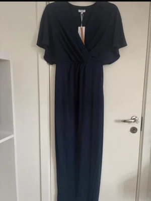 Marinblå långklänning från Bubbleroom - Snygg marinblå långklänning från Bubbleroom med omlott-design och v-ringad hals. Klänningen har korta, fladdriga ärmar och markerad midja. Perfekt för dig som vill ha en elegant och stilren look. Materialet är mjukt och består till stor del av återvunnen polyester.