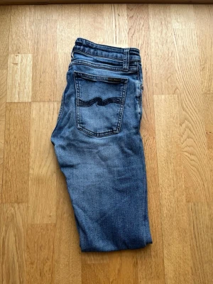  Nudie Jeans - Snygga blå slim fit jeans från Nudie Jeans med klassisk femficksmodell och tydliga slitningar framtill. Jeansen har en mörkblå broderad detalj på bakfickan och är tillverkade i mjuk denim. Perfekta för en avslappnad och trendig look.