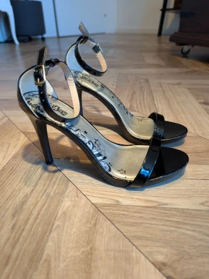 Svarta lackpumps med ankelrem - Snygga svarta pumps från Graceland i lackmaterial med smal rem över tårna och justerbar ankelrem med spänne. Klackarna är höga och smala, och innersulan har ett dekorativt mönster med stjärnor. Perfekta för att lyfta din outfit.