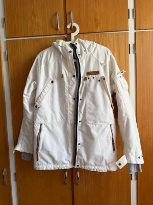 Vit vindjacka Peak Performance S - Vit Skidjacka från Peak Performance med flera fickor, dragkedja och tryckknappar. Jackan har huva, detaljer i brunt och grått innerfoder. Perfekt för blåsiga dagar och har en sportig, stilren look.