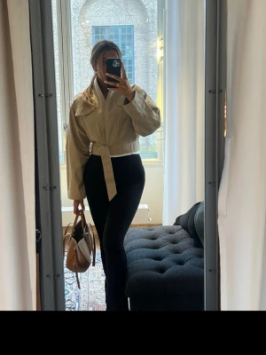 Stilren brun handväska i läder - Säljer en beige kort jacka med bälte i midjan och stora kragar. Jackan har en modern och boxig passform, långa ärmar och är tillverkad i ett slätt material som känns lätt och bekvämt. Perfekt för dig som gillar stilrena och trendiga plagg. Nypris 999, s/xs