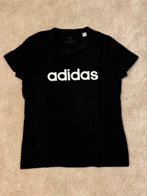 Svart Adidas t-shirt med logga - Snygg svart t-shirt från Adidas med stor vit logga framtill. Klassisk rund halsringning och korta ärmar. Tillverkad i mjuk bomull som känns skön mot huden. Perfekt för en sportig och avslappnad stil.
