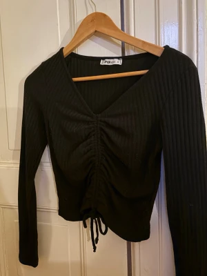 Svart ribbad omlottopp med snörning - Säljer en svart ribbad topp från FBsisermed v-ringning och snörning framtill. Toppen är croppad men går att ändra med snören och har långa ärmar. Perfekt för dig som gillar en stilren och trendig look.