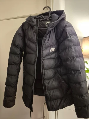 Svart pufferjacka från Nike med huva - Säljer en svart pufferjacka från Nike med huva och dragkedja framtill. Jackan har två sidofickor med knapp och en broderad Nike-logga på bröstet. Perfekt för kalla dagar och riktigt skön att bära. Klassisk pufferdesign med quiltade sektioner.