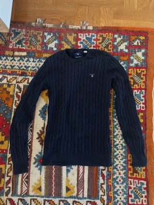 Mörkblå kabelstickad tröja GANT - Kabelstickad mörkblå tröja från GANT i stretchig bomull. Tröjan har rund halsringning, långa ärmar och diskret GANT-logga broderad på bröstet. Perfekt för en stilren och klassisk look.