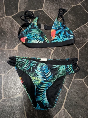 Tropisk bikini med papegojor stl 13-15 år - Snygg bikini med tropiskt mönster i grönt, blått och inslag av rött och gult. Både överdel och underdel har svarta detaljer och flera smala band i sidorna. Bikinin har papegojor och palmblad på svart botten, vilket ger en färgstark och somrig look.