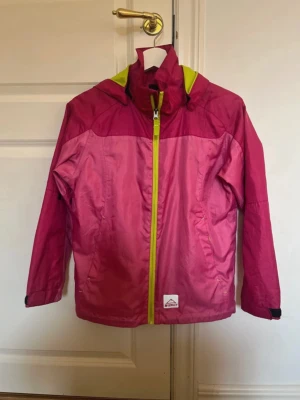 Rosa vindjacka med limegröna detaljer - Snygg rosa vindjacka från McKinley med limegröna detaljer på dragkedja och huva. Jackan har två fickor med dragkedja, justerbara ärmslut och en praktisk huva. Perfekt för blåsiga dagar och aktiviteter.
