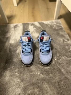 Jordan 4 University Blue sneakers - Säljer ett par Jordan 4 University Blue sneakers i ljusblå mocka med meshdetaljer och grå sula. Skorna har svarta och vita inslag samt klassisk Jumpman-logga på plösen. Snygga detaljer och ikonisk siluett som sticker ut.