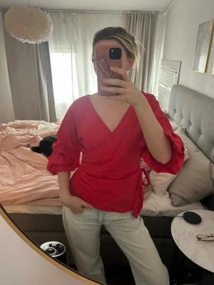 Röd omlottblus med volangärmar - Snygg röd omlottblus med trekvartsärm och volangdetaljer. Blusen har knytband i midjan och djup v-ringning. Perfekt för dig som vill ha en färgstark och trendig look. Materialet ser ut att vara lätt och bekvämt.