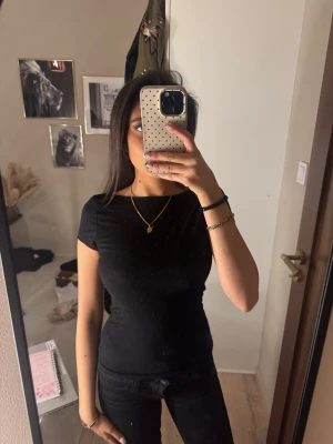 Svart topp med öppenrygg - En enkel svart t-shirt med rund hals och korta ärmar. Snygg passform som sitter tight mot kroppen, perfekt att matcha med jeans eller kjol. Materialet känns mjukt och bekvämt, troligen bomull eller polyester. Inga synliga loggor eller mönster.