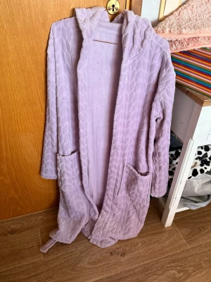 Ljuslila morgonrock med huva - Mysig ljuslila morgonrock i mjukt fleece-material med huva och knytskärp i midjan. Har två stora fickor framtill och ett diskret mönster i tyget. Perfekt för att chilla hemma och hålla sig varm. Storleken är onesize, mej passar den bra och jag har storlek 48