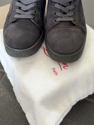 Gråa sneakers från Christian Louboutin - Snygga gråa sneakers från Christian Louboutin i mocka med klassisk låg profil och matchande gråa skosnören. Skorna har en stilren design med diskreta sömmar och en robust gummisula. Perfekt för dig som gillar exklusiva och trendiga skor.