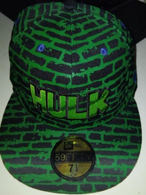 Grön Hulk keps från New Era - Säljer en grön och svart 59FIFTY keps från New Era med Hulk-tryck framtill. Kepsen har ett mönster som liknar tegelväggar och broderad text i grönt och svart. Märkeslogga på sidan och officiella klistermärken kvar på skärmen.