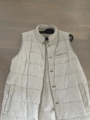 Beige dunväst från John Henric - Stilren beige dunväst från John Henric med ståkrage och knäppning framtill. Västen har två sidofickor och en bröstficka, quiltad design och är tillverkad i ett mjukt material med dunfyllning. Perfekt för lager på lager och en clean look. Köpt för 1700 