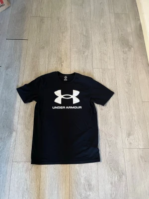  Under Armour S - Svart t-shirt från Under Armour med stort vitt logotryck på bröstet. Klassisk rund hals och korta ärmar. Perfekt för en sportig stil och tillverkad i ett lätt och bekvämt material.