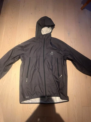 Svart regnjacka från Haglöfs - Snygg svart regnjacka från Haglöfs med huva och dragkedja framtill. Jackan har två stora fickor med dragkedja och diskret logga på bröstet. Tillverkad i vattentätt material, perfekt för regniga dagar. Enkel och stilren design. Är en XL men passar mer som en M eller liten L. 