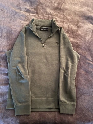 Olivgrön half-zip tröja Abercrombie - Snygg olivgrön tröja från Abercrombie & Fitch med half-zip och ståkrage. Tröjan har diskret logga på bröstet och ribbade muddar vid ärmslut och nederkant. Perfekt för lager på lager och enkel att matcha med jeansen.