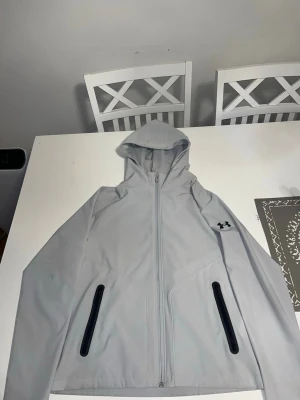 Ljusgrå vindjacka från Under Armour - Snygg ljusgrå vindjacka från Under Armour med huva och hel dragkedja framtill. Jackan har två svarta dragkedjeförsedda fickor och meshfoder på insidan för extra komfort. Perfekt för blåsiga dagar och sportiga aktiviteter.