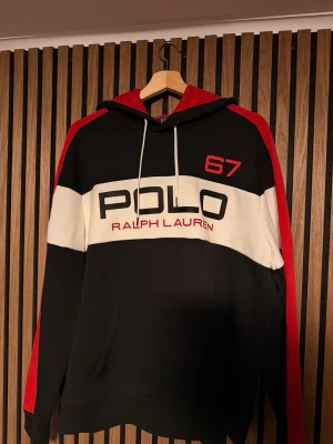 Svart och röd hoodie från Polo Ralph Lauren - Snygg hoodie från Polo Ralph Lauren i svart, vitt och rött med stor logga och texten 'POLO 67' framtill. Hoodien har huva med dragsko, känguruficka och breda färgblock. Perfekt för en sportig och avslappnad stil.