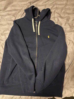 Mörkblå hoodie från Polo Ralph Lauren - Mörkblå hoodie med dragkedja från Polo Ralph Lauren. Klassisk design med huva och vita snören, samt gul broderad logga på bröstet. Mjuk bomullskvalitet och fickor framtill. Perfekt för en avslappnad och stilren look.