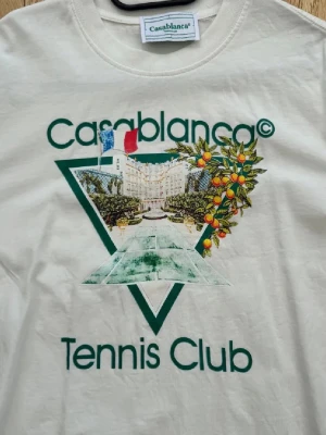 Vit sweatshirt Casablanca Tennis Club - Vit sweatshirt från Casablanca med tryck framtill där det står 'Casablanca Tennis Club' och ett färgglatt motiv med frukter och flaggor. Tröjan har rund halsringning och ribbade muddar. Perfekt för dig som gillar sportig och trendig stil.