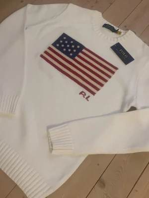 Ralph Lauren Tröja  - Vit stickad tröja från Polo Ralph Lauren med amerikansk flagga i rött, blått och vitt på bröstet. Tröjan har rund halsringning, ribbstickade muddar och nederkant samt RL-broderi under flaggan. Perfekt för dig som gillar klassisk och stilren design.