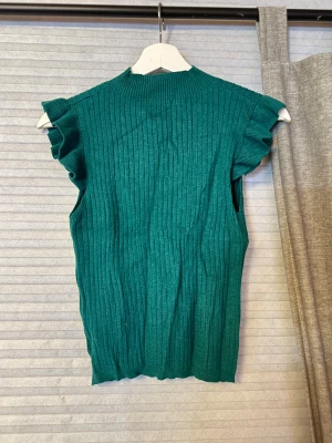 Grön ribbad ärmlös topp med volang - Snygg grön ribbad topp med volangdetaljer vid axlarna. Toppen är ärmlös och har en hög halsringning. Perfekt att styla med jeans eller kjol för en trendig look.