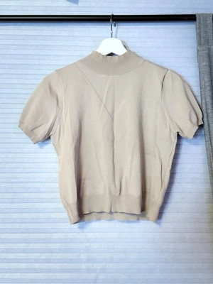 Beige ribbad topp med kort ärm - Snygg beige topp med ribbad hög krage och korta ärmar. Perfekt till jeans eller kjol för en clean och stilren look. Materialet känns mjukt och stretchigt.