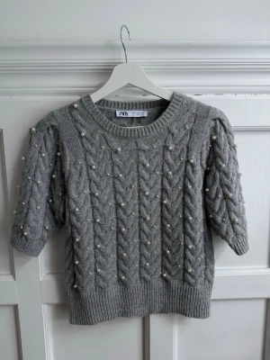 Grå stickad tröja med pärlor från Zara - Supersöt grå stickad tröja från Zara med korta puffärmar och dekorativa pärlor. Tröjan har ett klassiskt flätmönster och ribbade muddar vid ärmslut och nederkant. Tröjan är superskön, sticka inte och passar perfekt till både varma och kalla dagar 🤗 