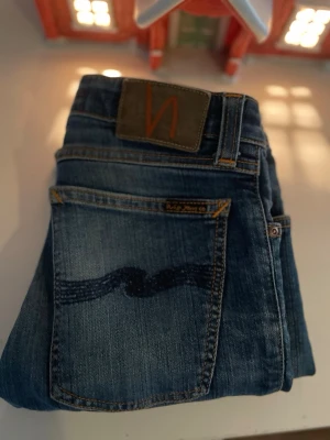 Blå jeans från Nudie Jeans - Snygga blå jeans från Nudie Jeans med klassisk femficksdesign och tydlig vågdetalj på bakfickan. Jeansen har en rak passform och är gjorda i denim med orangea sömmar och läderpatch i midjan. Stora i storleken !