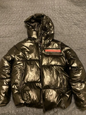 Svart pufferjacka från Prada - Säljer en svart, glansig pufferjacka från Prada med huva och flera dragkedjeförsedda fickor. Jackan har en röd detalj med Prada-logga på bröstet och är tillverkad i polyamid med graphene padding. Perfekt för kalla dagar och har en riktigt modern vibe.