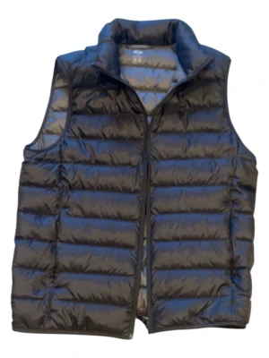 Svart dunväst från Uniqlo - Stilren svart dunväst från Uniqlo med hög krage och quiltad design. Västen har dragkedja framtill och är lätt vadderad för extra värme. Perfekt att slänga över en hoodie eller tröja när det är kyligt ute.